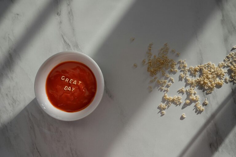 A flatlay of tomato soup with 'Great Day' spelled in pasta on a marble background.